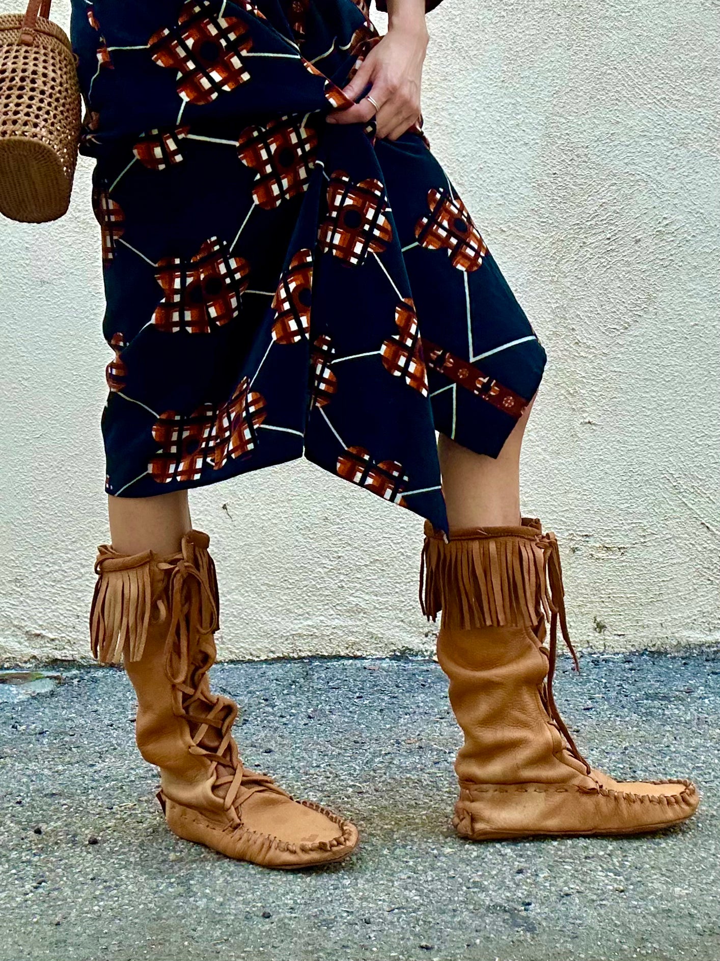 Person wearing a patterned dress and brown fringed boots against a light-colored wall.