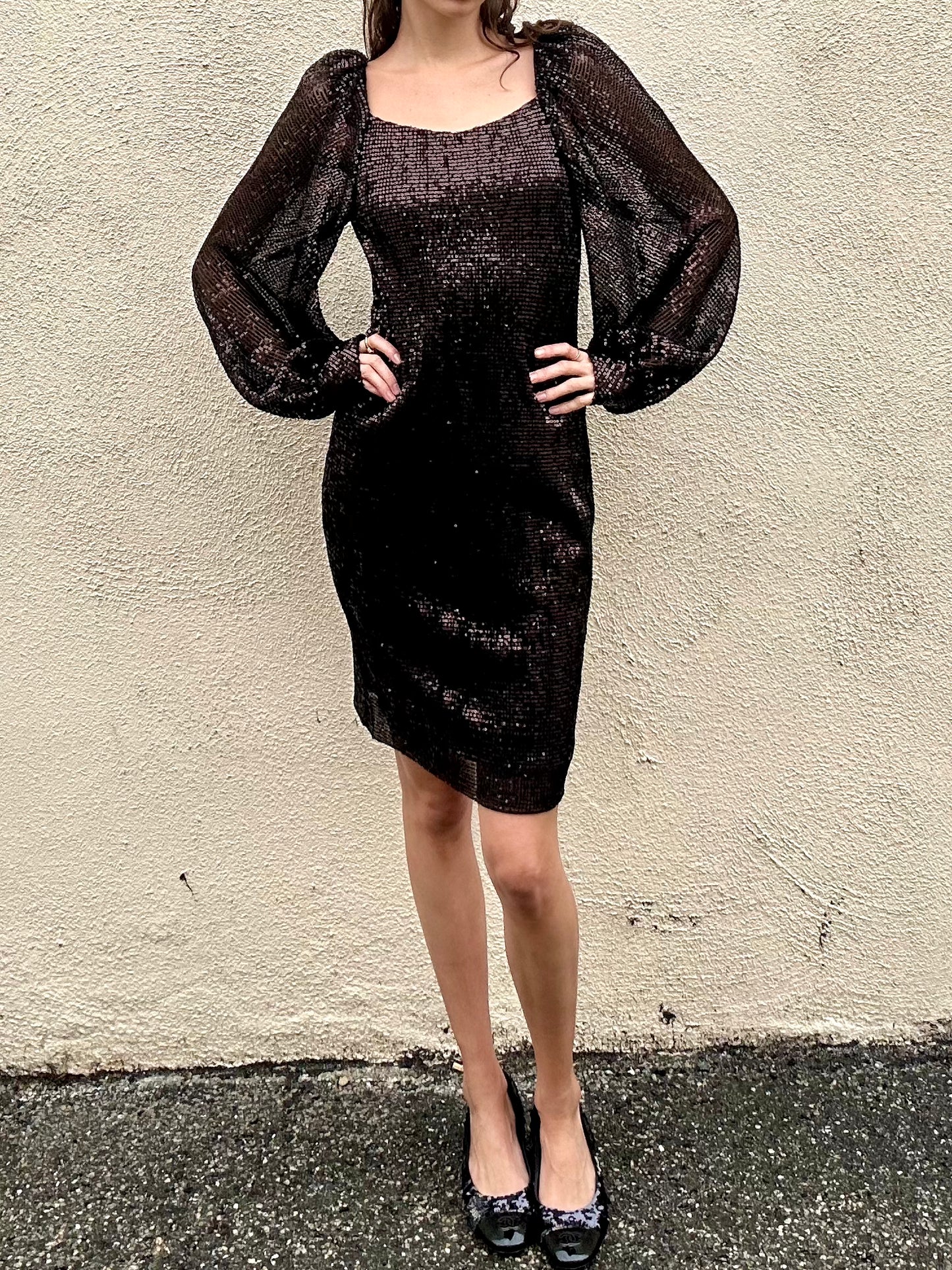 Person wearing a black dress with sheer sleeves against a beige wall.